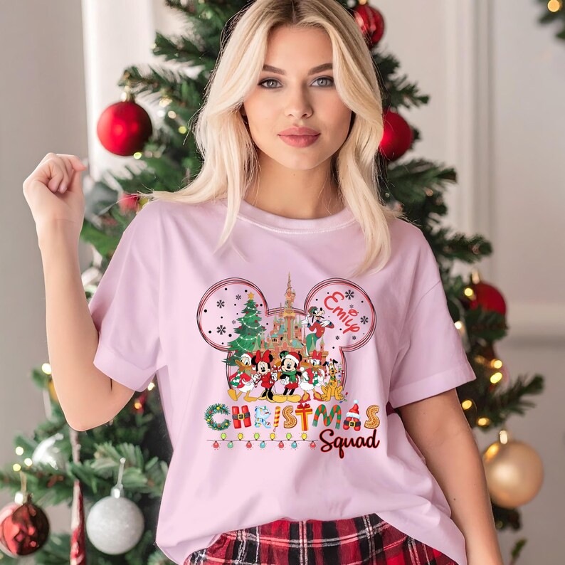 May include: A light pink t-shirt featuring a Mickey Mouse-themed Christmas design. The design includes Mickey and Minnie Mouse, a Christmas tree, and the words "Christmas Squad". The name "Emily" is also included in the design.