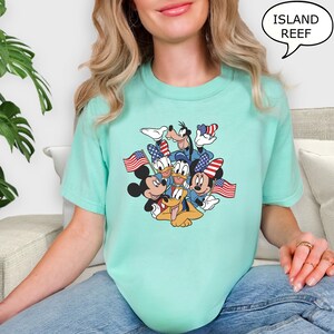 May include: A light teal t-shirt featuring a graphic of cartoon characters, including Mickey Mouse, Donald Duck, and Goofy, holding American flags. The text "ISLAND REEF" is in a speech bubble. The shirt is made of a soft material.