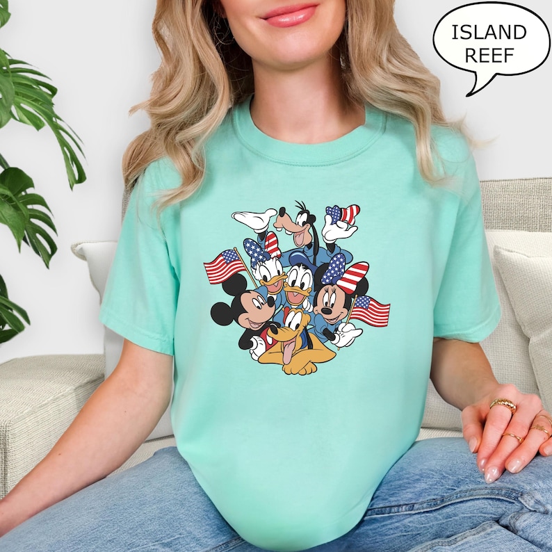 May include: A light teal t-shirt featuring a graphic of cartoon characters, including Mickey Mouse, Donald Duck, and Goofy, holding American flags. The text "ISLAND REEF" is in a speech bubble. The shirt is made of a soft material.