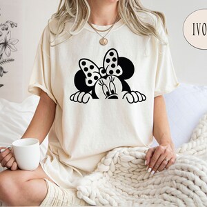 K&ouml;nnte beinhalten: Ein wei&szlig;es T-Shirt mit einer schwarz-wei&szlig;en Illustration von Minnie Mouse, die &uuml;ber eine Oberfl&auml;che schaut. Die Illustration zeigt Minnies charakteristisches gepunktetes Band.