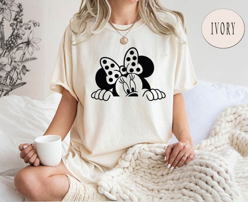K&ouml;nnte beinhalten: Ein wei&szlig;es T-Shirt mit einer schwarz-wei&szlig;en Illustration von Minnie Mouse, die &uuml;ber eine Oberfl&auml;che schaut. Die Illustration zeigt Minnies charakteristisches gepunktetes Band.