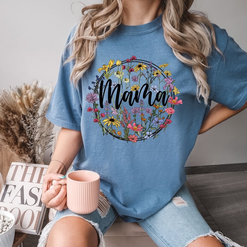 Peut inclure: Un t-shirt bleu clair avec un motif circulaire floral et le mot "Mama" en texte noir.