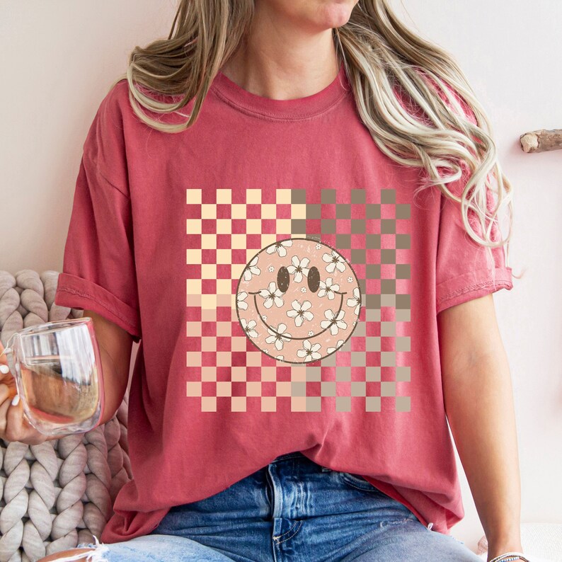 Peut inclure: Un t-shirt rouge avec un motif &agrave; carreaux et un smiley rose avec des fleurs blanches. Le smiley est centr&eacute; sur le t-shirt.