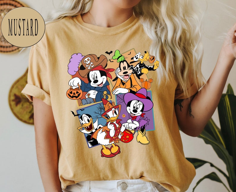 K&ouml;nnte beinhalten: Ein senfgelbes T-Shirt mit einem grafischen Druck von Mickey Mouse, Minnie Mouse, Donald Duck, Goofy und Pluto in Halloween-Kost&uuml;men. Die Figuren halten K&uuml;rbisse und S&uuml;&szlig;igkeiten.