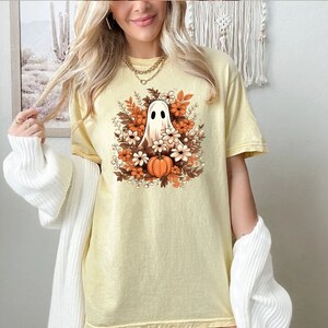 Op de afbeelding: Een geel T-shirt met een witte geest omringd door oranje en witte bloemen en een kleine oranje pompoen. De geest heeft een klein gezicht met twee ogen.