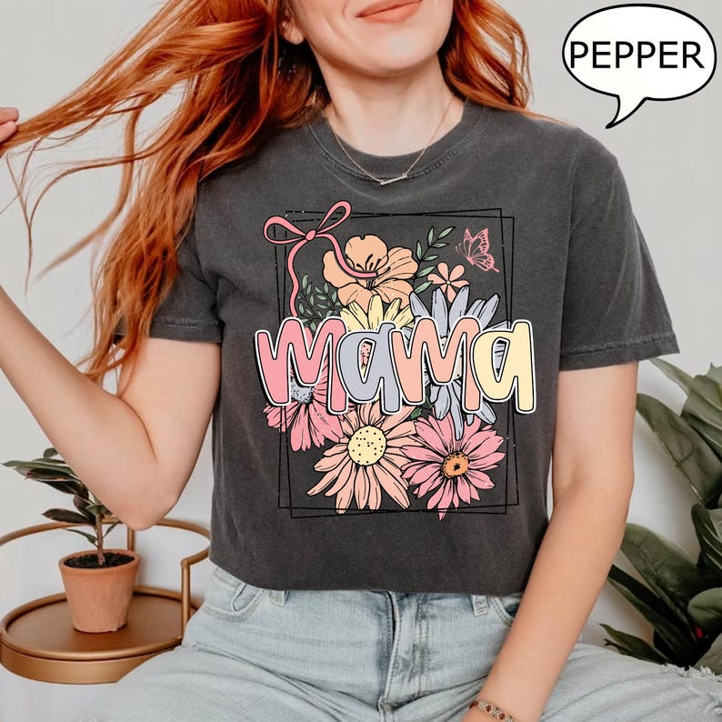 May include: A dark grey t-shirt with a floral design and the word "mama" in pink, yellow, and blue lettering. The design is framed in a black rectangle.