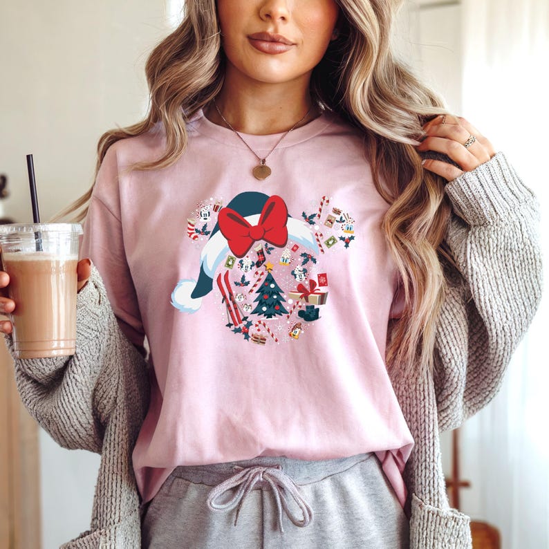 Puede incluir: Una camiseta de manga larga rosa claro con un dise&ntilde;o navide&ntilde;o. El dise&ntilde;o incluye un gorro de Pap&aacute; Noel con un lazo rojo, un &aacute;rbol de Navidad, esqu&iacute;s y cajas de regalo. El dise&ntilde;o est&aacute; rodeado de bastones de caramelo y otros elementos festivos.