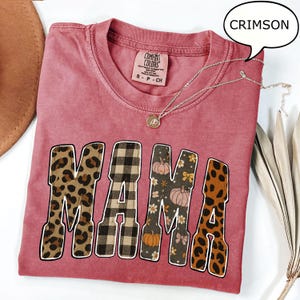 May include: Crimson-colored t-shirt with the word "MAMA" in a bold, decorative font. The letters feature leopard print, plaid, and floral patterns. A silver necklace with a pendant rests on the shirt.
