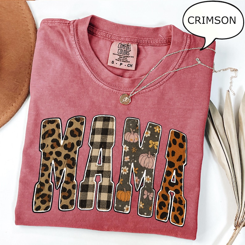 May include: Crimson-colored t-shirt with the word "MAMA" in a bold, decorative font. The letters feature leopard print, plaid, and floral patterns. A silver necklace with a pendant rests on the shirt.