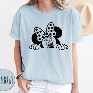 K&ouml;nnte beinhalten: Ein hellblaues T-Shirt mit einem schwarz-wei&szlig;en Motiv von Minnie Mouse, die &uuml;ber den Rand schaut. Minnie Mouse tr&auml;gt eine rote Schleife mit wei&szlig;en Punkten.