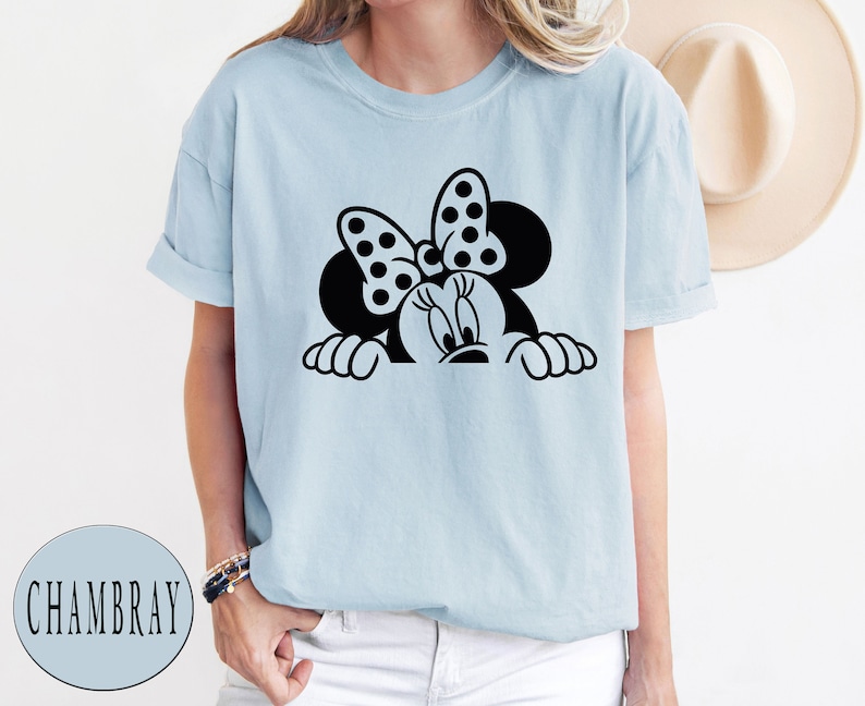 K&ouml;nnte beinhalten: Ein hellblaues T-Shirt mit einem schwarz-wei&szlig;en Motiv von Minnie Mouse, die &uuml;ber den Rand schaut. Minnie Mouse tr&auml;gt eine rote Schleife mit wei&szlig;en Punkten.