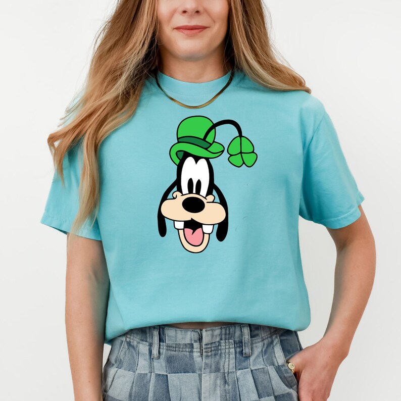 Puede incluir: Una camiseta azul claro con un personaje de dibujos animados con un sombrero verde y un tr&eacute;bol de cuatro hojas. El personaje tiene una cara en blanco y negro con la boca abierta. La camiseta es de manga corta y de material suave.