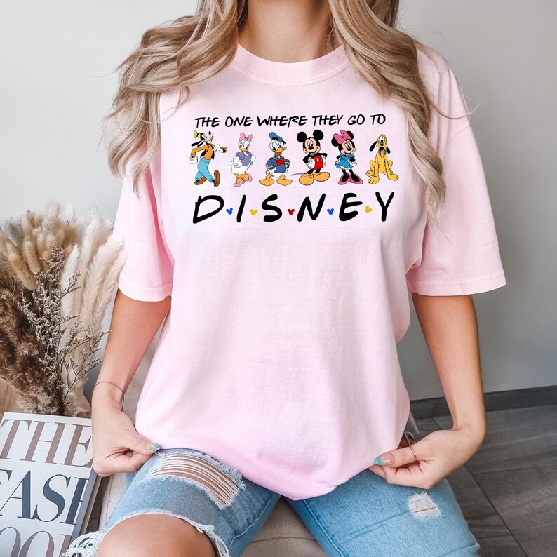 May include: A light pink t-shirt featuring the text "THE ONE WHERE THEY GO TO DISNEY" above a row of cartoon characters, including Goofy, Donald Duck, Daisy Duck, Mickey Mouse, Minnie Mouse, and Pluto. The word "DISNEY" is printed below.