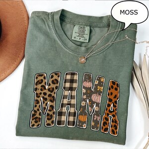May include: A moss green t-shirt with the word "MAMA" in a mix of patterns, including leopard print, plaid, and floral designs. A silver necklace with a pendant is draped across the shirt. The shirt is a Comfort Colors brand.