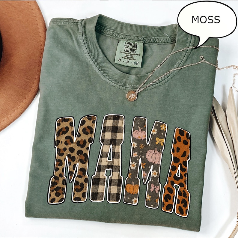 May include: A moss green t-shirt with the word "MAMA" in a mix of patterns, including leopard print, plaid, and floral designs. A silver necklace with a pendant is draped across the shirt. The shirt is a Comfort Colors brand.