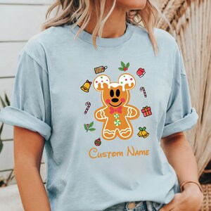 May include: Light blue t-shirt with a gingerbread Mickey Mouse design. The design includes a gingerbread Mickey Mouse with red bow tie, surrounded by candy canes, bells, and holly. The text "Custom Name" is printed below the design.
