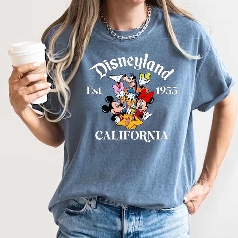 Puede incluir: Una camiseta azul con un gr&aacute;fico blanco de Mickey Mouse y sus amigos. El gr&aacute;fico incluye el texto "Disneyland Est 1955 California".