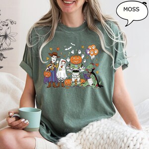 Puede incluir: Camiseta verde musgo con un gr&aacute;fico de dibujos animados de Halloween. El dise&ntilde;o muestra personajes disfrazados, como Woody, Buzz Lightyear y un dinosaurio leyendo. La palabra "MOSS" est&aacute; en un bocadillo.