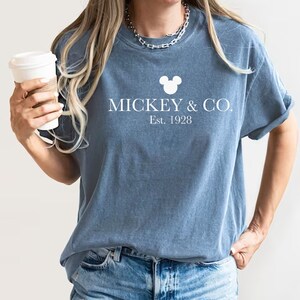 Puede incluir: Una camiseta azul con una cabeza de Mickey Mouse blanca y el texto "MICKEY & CO. Est. 1928"