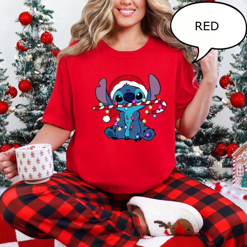 Puede incluir: Una camiseta roja de manga corta con un personaje de dibujos animados que lleva un gorro de Pap&aacute; Noel y sostiene un bast&oacute;n de caramelo. El personaje es azul con una guirnalda de luces. La palabra "RED" est&aacute; en un bocadillo. La persona lleva un pijama a cuadros rojos y negros.
