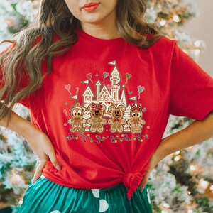 May include: Red t-shirt featuring a gingerbread castle and gingerbread characters. The design includes a castle, gingerbread people, and festive lights. The shirt is a solid red color and has a crew neck.