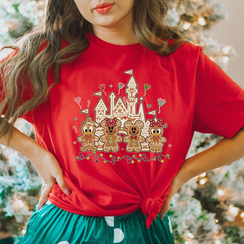 May include: Red t-shirt featuring a gingerbread castle and gingerbread characters. The design includes a castle, gingerbread people, and festive lights. The shirt is a solid red color and has a crew neck.