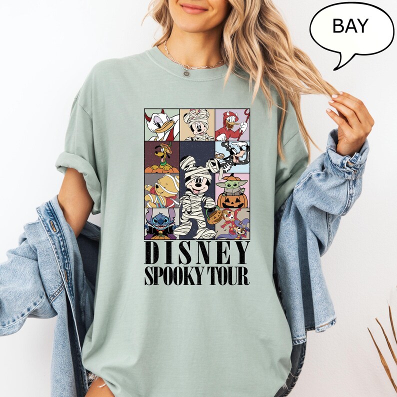 May include: A light green t-shirt featuring a grid of Disney characters in Halloween costumes. The shirt has the words "DISNEY SPOOKY TOUR" printed in black. The shirt is a relaxed fit.