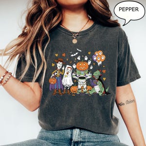 Puede incluir: Camiseta gris oscuro con un gr&aacute;fico de Halloween con personajes de dibujos animados disfrazados. El dise&ntilde;o incluye a Woody, un fantasma, Buzz Lightyear y un dinosaurio, con calabazas y fantasmas. La palabra "PEPPER" est&aacute; en una burbuja.