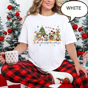 May include: White t-shirt featuring a festive graphic of Disney characters, including Mickey and Minnie Mouse, in holiday attire. The shirt has the text "It's the most wonderful time of the year". The person is wearing red and black plaid pajama pants.
