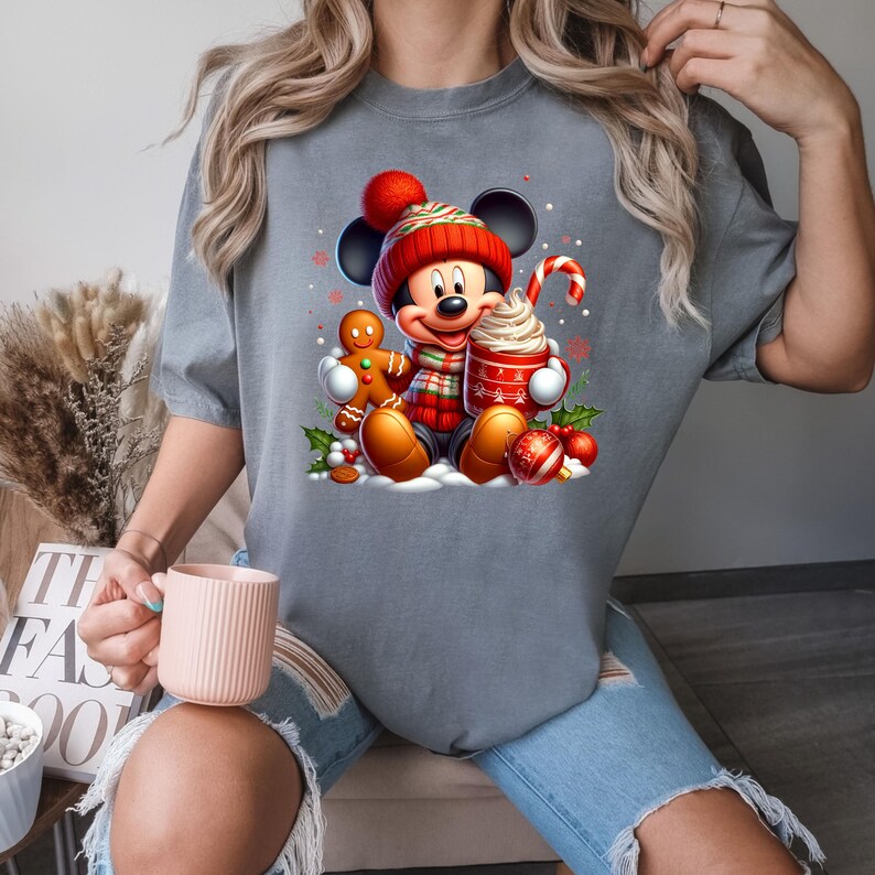 Puede incluir: Una camiseta gris con un gr&aacute;fico navide&ntilde;o con Mickey Mouse con un gorro de Pap&aacute; Noel rojo, sosteniendo una taza de chocolate caliente y un hombre de jengibre.