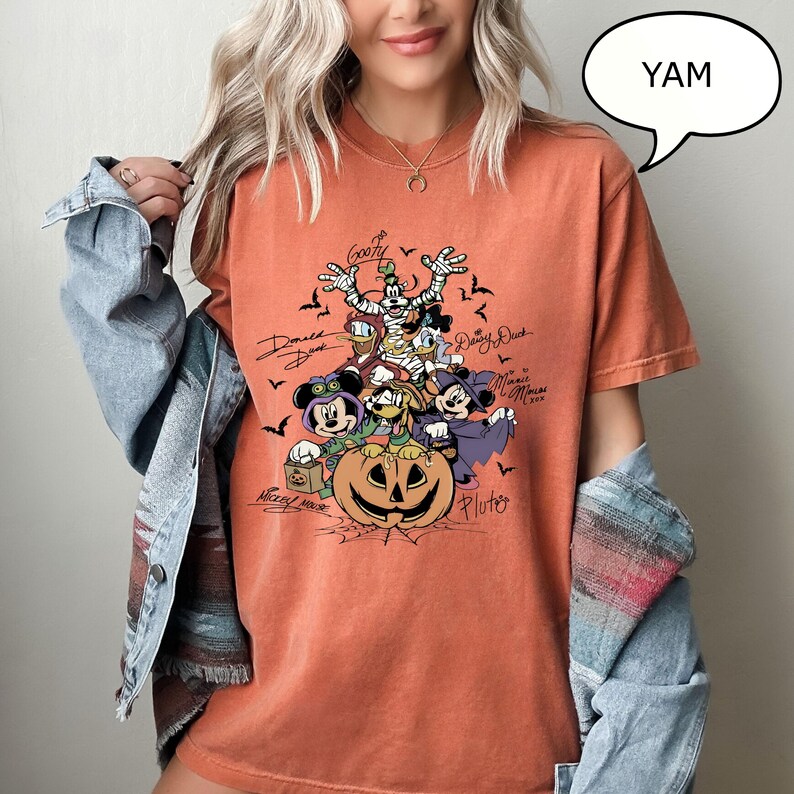 May include: Orange t-shirt featuring a Halloween-themed graphic with Disney characters, including Mickey Mouse, Donald Duck, and Goofy, surrounding a carved pumpkin. The shirt has character signatures and the word "YAM" in a speech bubble.
