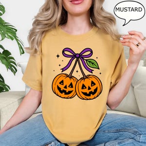 May include: Mustard-colored t-shirt featuring a Halloween-themed graphic. The graphic depicts two orange jack-o'-lanterns connected by a purple bow and green leaf. The word "MUSTARD" is in a speech bubble. The shirt is a casual wear item.