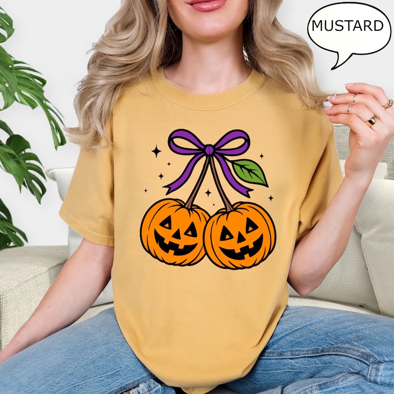 May include: Mustard-colored t-shirt featuring a Halloween-themed graphic. The graphic depicts two orange jack-o'-lanterns connected by a purple bow and green leaf. The word "MUSTARD" is in a speech bubble. The shirt is a casual wear item.