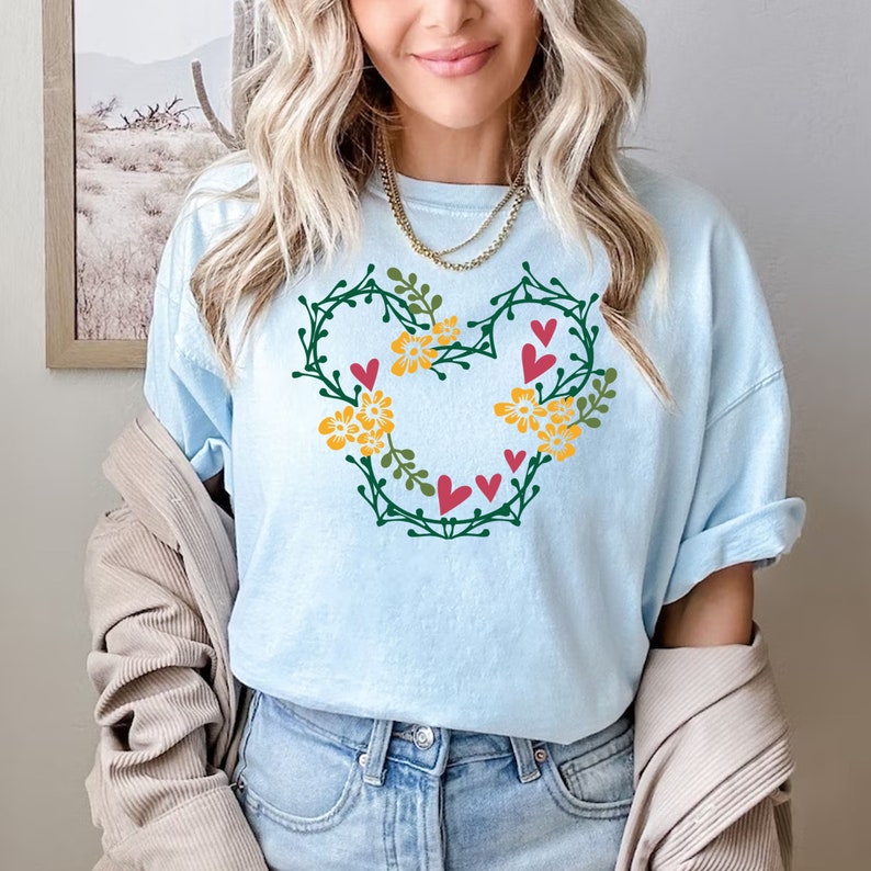 K&ouml;nnte beinhalten: Ein hellblaues T-Shirt mit einem gr&uuml;nen floralen Mickey-Maus-Design. Das Design enth&auml;lt rote Herzen und gelbe Blumen.