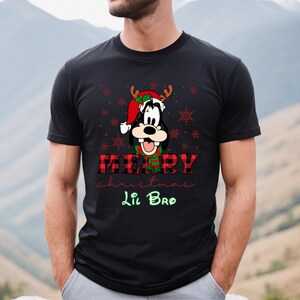 Op de afbeelding: Zwart T-shirt met een cartoon afbeelding van Goofy met een kerstmanmuts en rendiergewei. De tekst "Merry Christmas Lil Bro" staat op het shirt gedrukt.