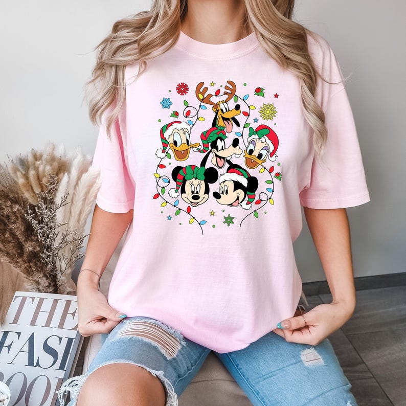 May include: Pink t-shirt with a Christmas-themed design featuring Mickey Mouse, Minnie Mouse, Donald Duck, Goofy, and Pluto. The characters are wearing Santa hats and surrounded by Christmas lights.
