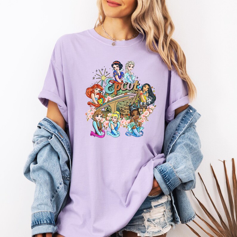 May include: A lavender t-shirt featuring a graphic of Disney princesses in front of the Epcot logo. The princesses are wearing their signature dresses and are surrounded by flowers.