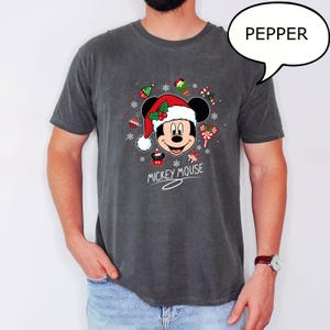 May include: Dark gray t-shirt featuring a festive Mickey Mouse design. Mickey is wearing a Santa hat and surrounded by Christmas ornaments and candy canes. The words "Mickey Mouse" are written below the graphic.
