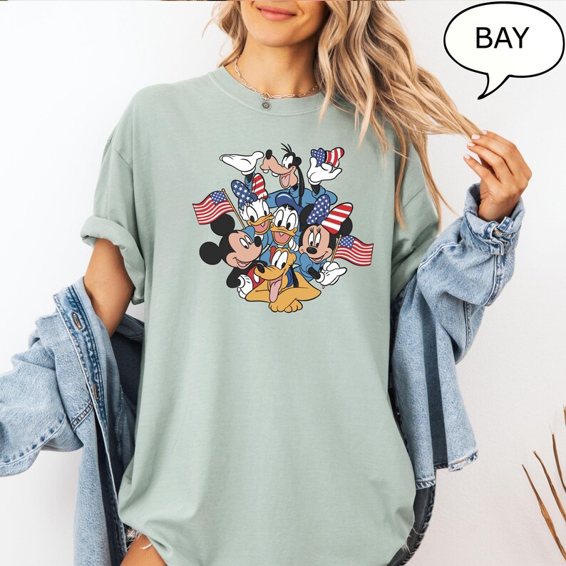 May include: A light green t-shirt featuring a graphic of Disney characters Mickey Mouse, Minnie Mouse, Donald Duck, Daisy Duck, Goofy, and Pluto. The characters are holding American flags and wearing patriotic accessories. The word "BAY" is in a speech bubble.