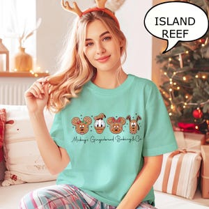 Puede incluir: Una camiseta de color verde azulado claro con dise&ntilde;os de personajes de pan de jengibre de Mickey, Donald, Minnie y Pluto. El texto "Mickey's Gingerbread Baking & Co." est&aacute; impreso debajo. Una burbuja de di&aacute;logo dice "ISLAND REEF".