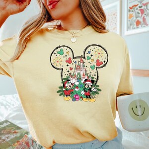 Puede incluir: Una camiseta amarilla con un gr&aacute;fico navide&ntilde;o con Mickey Mouse y Minnie Mouse frente al Castillo de Cenicienta. El gr&aacute;fico est&aacute; dentro de un contorno de oreja de Mickey Mouse con fuegos artificiales y corazones.