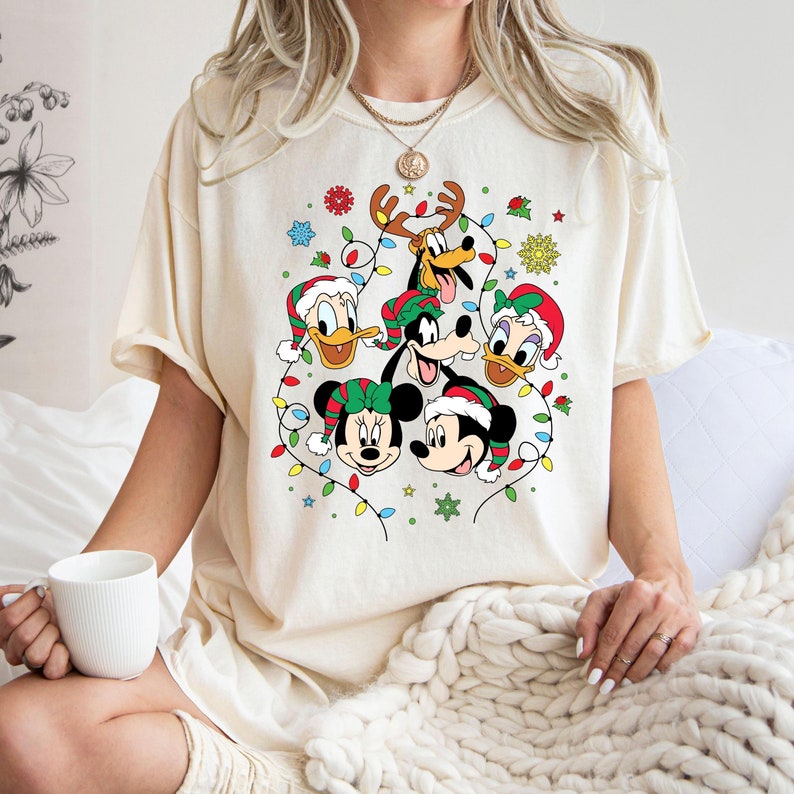 May include: A white t-shirt with a Christmas-themed design featuring Mickey Mouse, Minnie Mouse, Donald Duck, Goofy, and Pluto. The characters are surrounded by Christmas lights and snowflakes.