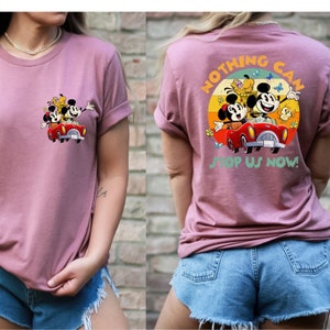 以下が含まれることがあります： ピンクのTシャツに、ミッキーマウス、ミニーマウス、プルートが赤い車に乗っているヴィンテージスタイルのグラフィックがプリントされています。グラフィックはシャツの後ろにあり、「Nothing Can Stop Us Now!」という文字が黄色と緑のフォントで書かれています。