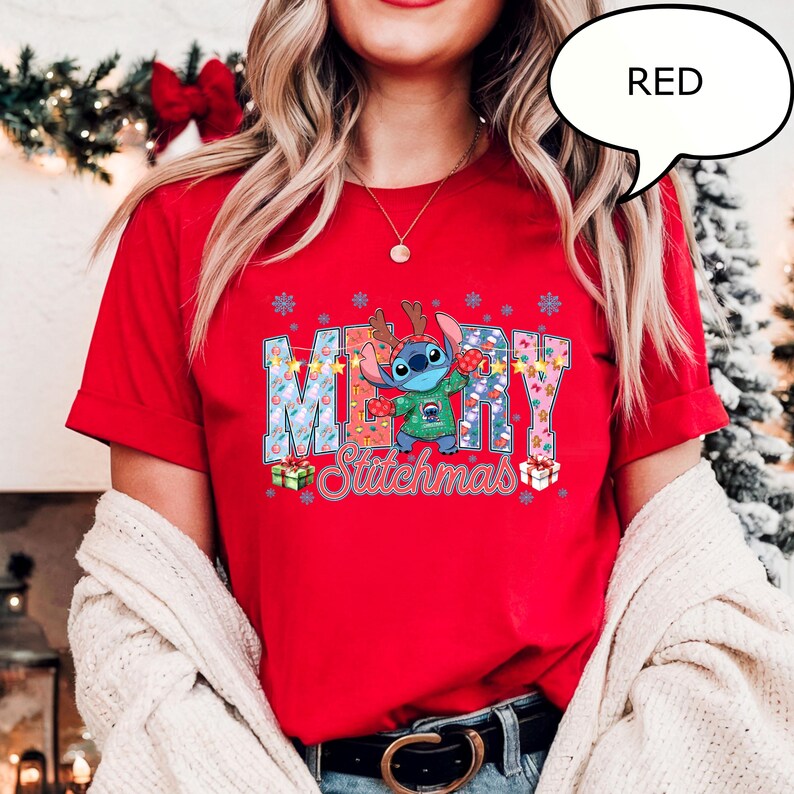 Op de afbeelding: Een rood t-shirt met het woord "MERRY" in een blauw en wit patroon, en een stripfiguur met een kersttrui. Het woord "Stitchmas" staat onder het personage. Een tekstballon zegt "RED".