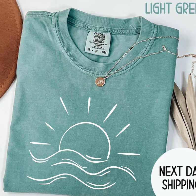 May include: A light green t-shirt with a white graphic of a sun setting over waves. The shirt has a tag that says "Comfort Colors" and "S - P - CH".
