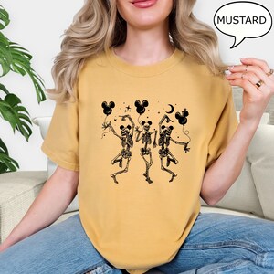 May include: Mustard-colored t-shirt featuring a black graphic of three dancing skeletons holding Mickey Mouse-shaped balloons. The word "MUSTARD" is in a speech bubble. The shirt is a casual, unisex style.