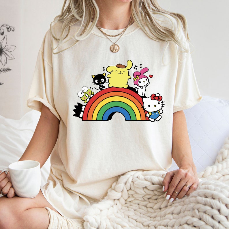 Pode incluir: Uma camiseta branca com um arco-&iacute;ris e personagens de desenhos animados. Os personagens incluem Hello Kitty, My Melody, Keroppi e um cachorro amarelo.