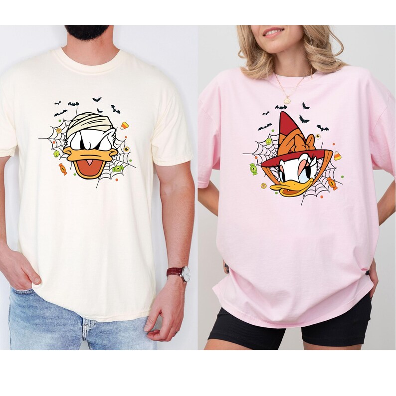 Może przedstawiać: Dwie koszulki, jedna kremowa i jedna r&oacute;żowa, z motywami kaczki z kresk&oacute;wek o tematyce Halloween. Kremowa koszulka ma kaczkę-mumię, a r&oacute;żowa koszulka przedstawia kaczkę-czarownicę. Obie koszulki mają pajęczyny, nietoperze i grafikę cukierk&oacute;w.