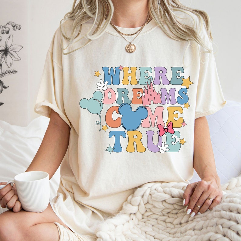 May include: A cream colored t-shirt with the text "Where Dreams Come True" in colorful letters. The text is surrounded by cartoon images of Mickey Mouse ears, a castle, stars, and balloons.