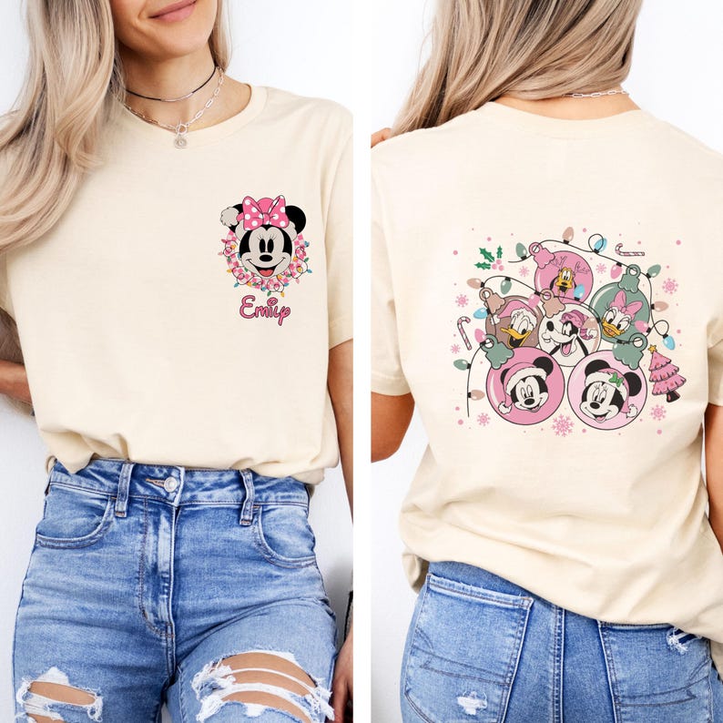 K&ouml;nnte beinhalten: Cremefarbenes T-Shirt mit einem Minnie Mouse-Motiv mit rosa Schleife und Blumenkranz sowie dem Namen "Emily". Die R&uuml;ckseite des Shirts zeigt Disney-Figuren in Weihnachtsbaumschmuck-Designs.